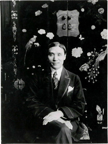 Photographie en noir et blanc de C. T. Loo, posant devant un meuble en laque à motif fleuri.