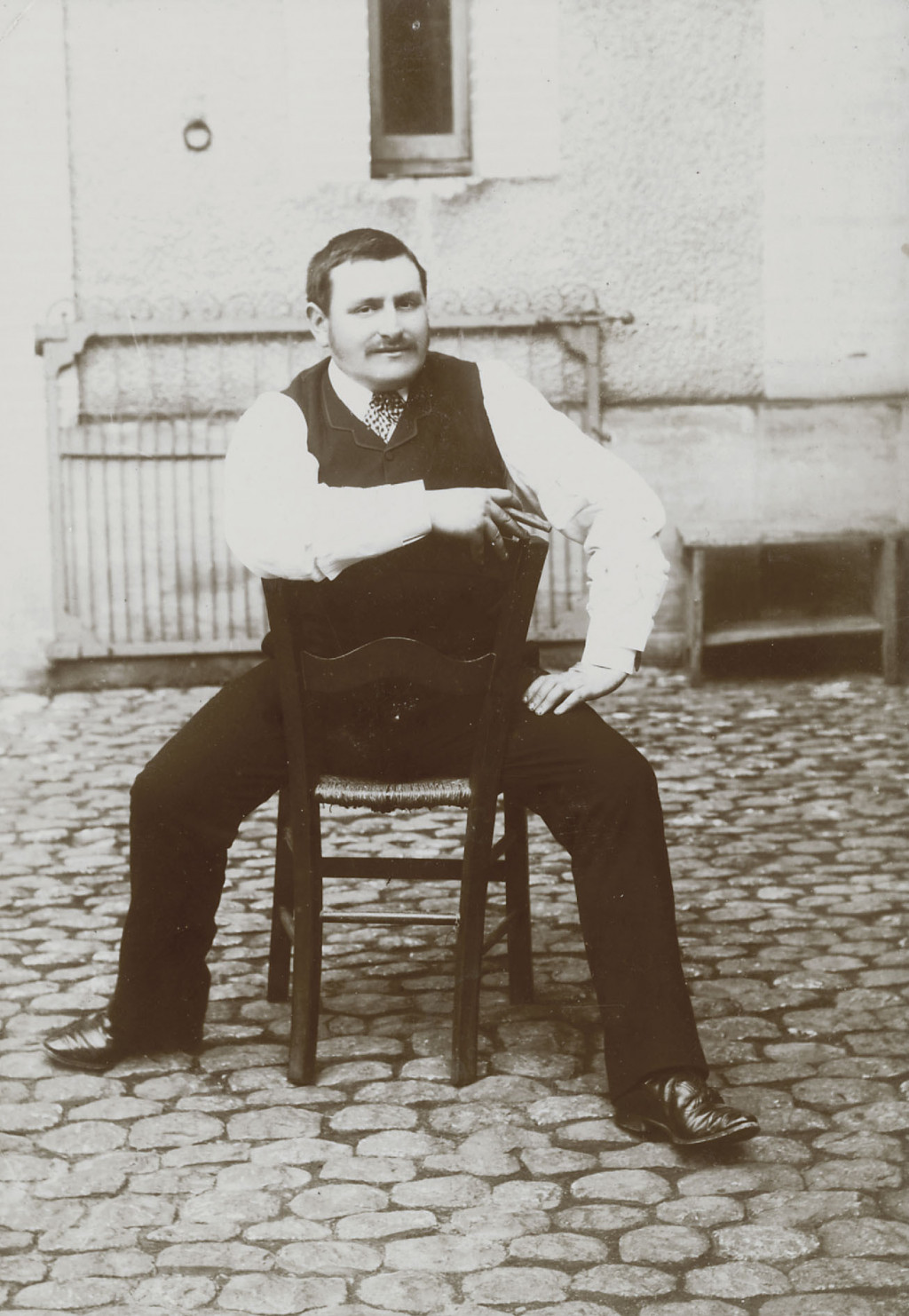 Photographie en noir et blanc de Georges Labit à califourchon sur une chaise, dans la rue.