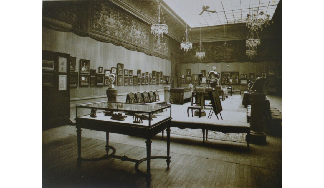 Black and white photograph of an auction room with paintings on the walls and objets in glass cases